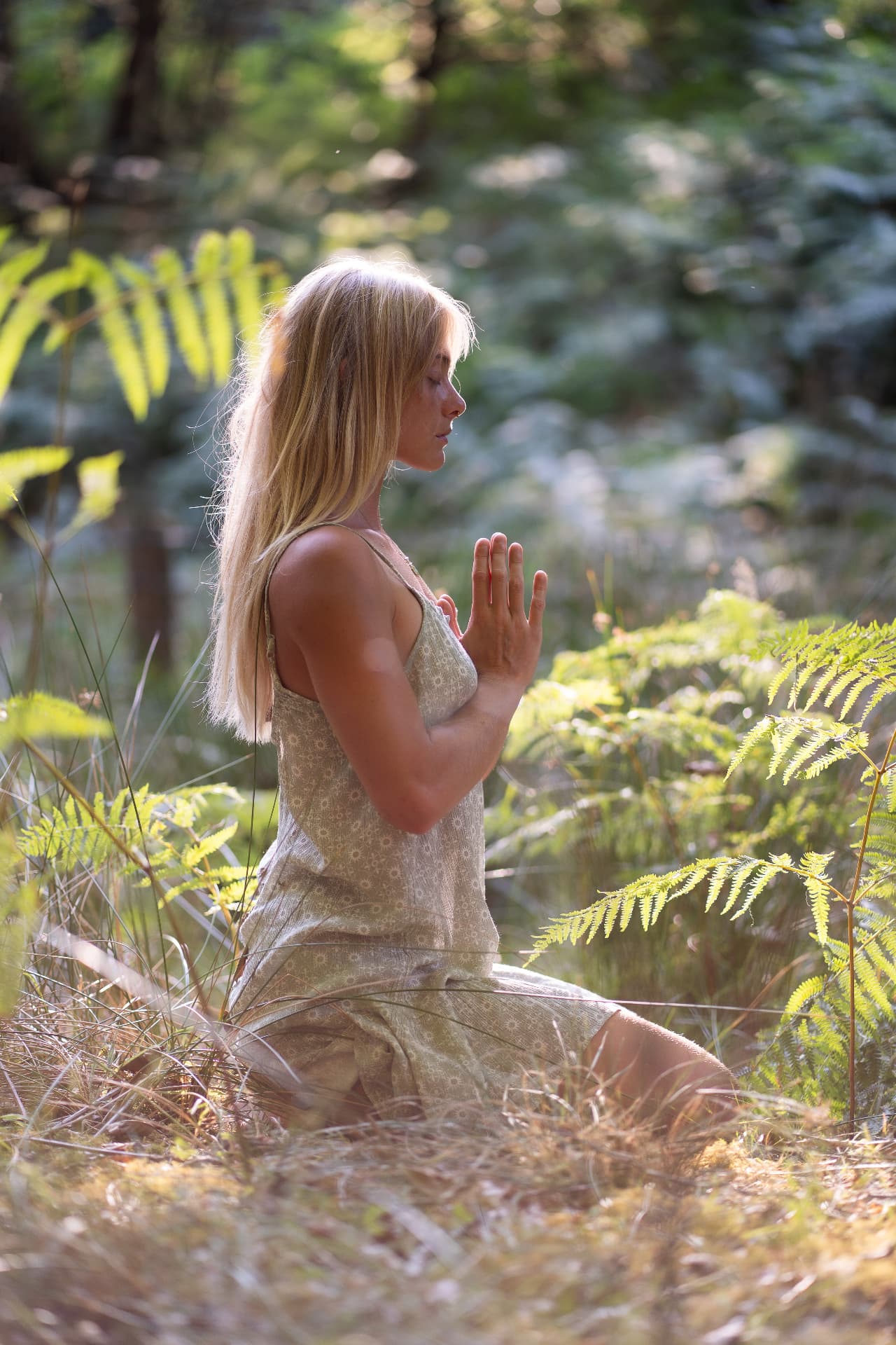 Woman in a sunlit woodland sitting among ferns with hands in a prayer gesture